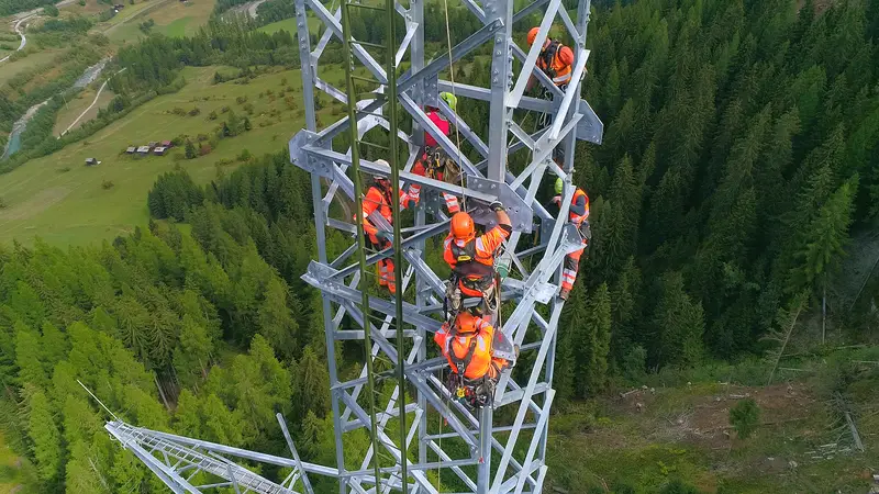 Monteure in orangefarbener Schutzkleidung arbeiten hoch oben an einem Freileitungsmast über einem Bergtal.