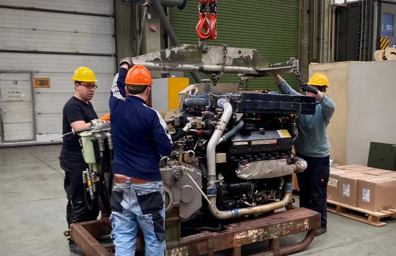 Drei Mechaniker mit Schutzhelmen arbeiten in einer Werkhalle an einem großen Motor am Kranhaken.