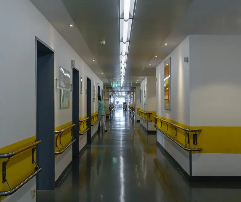 Ein langer Stationsflur mit gelben Handläufen, in der Tiefe eine Pflegekraft.