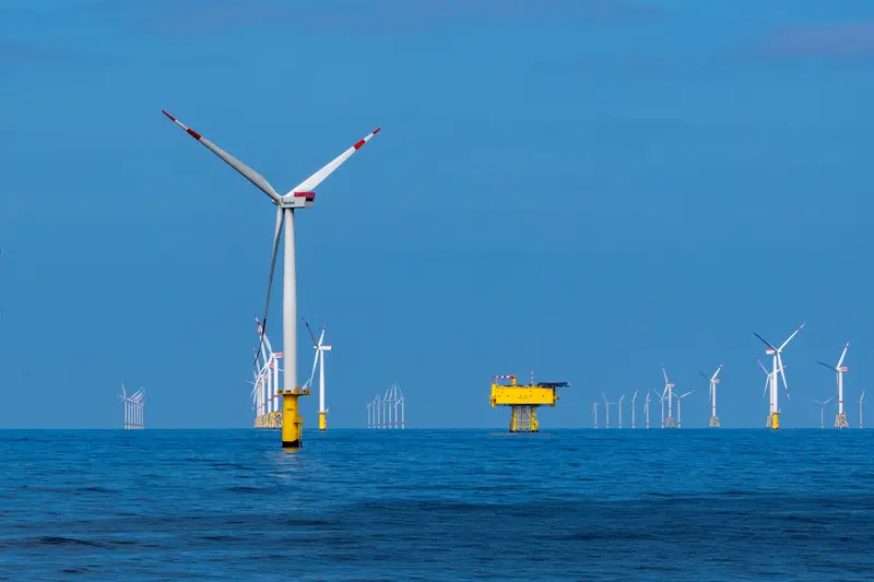 Windräder eines Offshore-Windparks auf offener See, dazwischen eine gelbe Umspannplattform.
